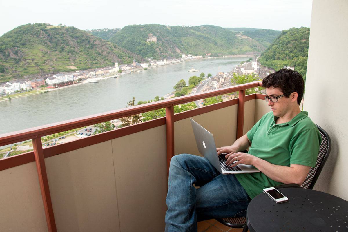 A photo of a man using a laptop while sitting on a balcony overlooking a river.