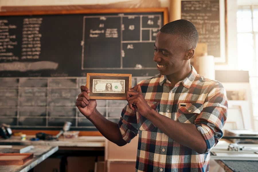 A photo of a man holding up the first dollar he made in a picture frame.