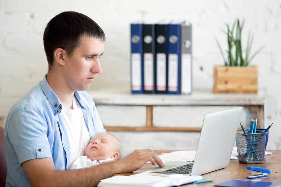 A photo of a man working from his home office while holding his baby.