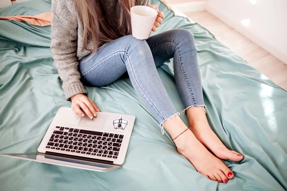 A photo of a woman starting her workday with coffee and a laptop on her bed at home.