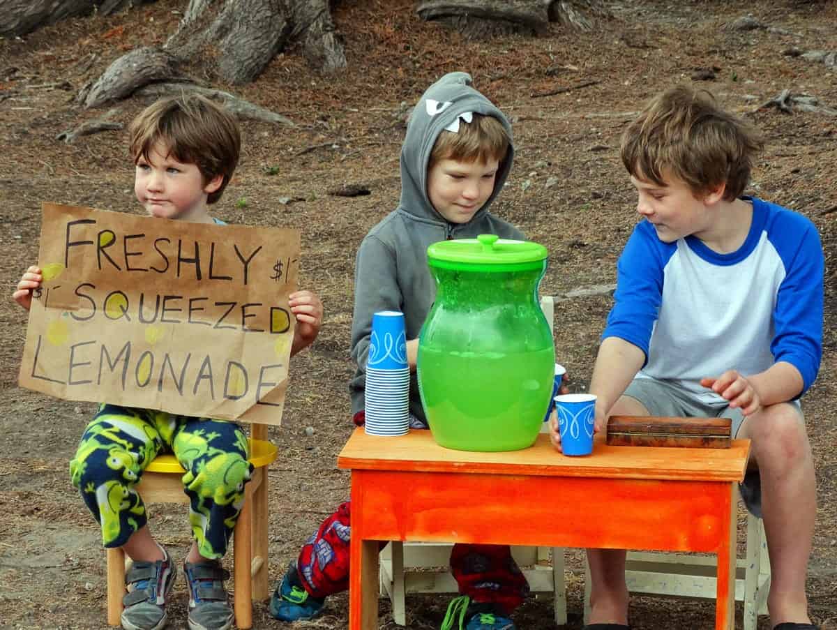 A photo of three young boys manning a lemonade stand in a park.