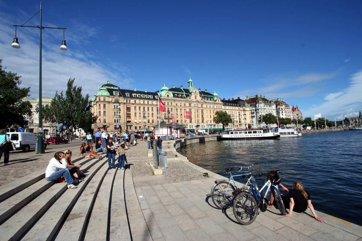 A photo of people sitting on concrete steps by a river running through a European city.