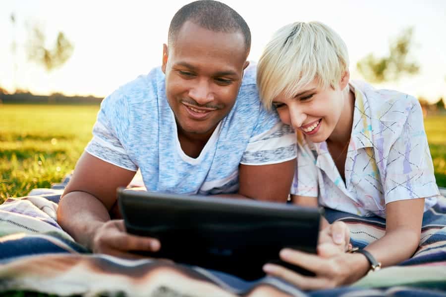 A photo of a couple lying on a blanket in the grass, looking at a tablet.