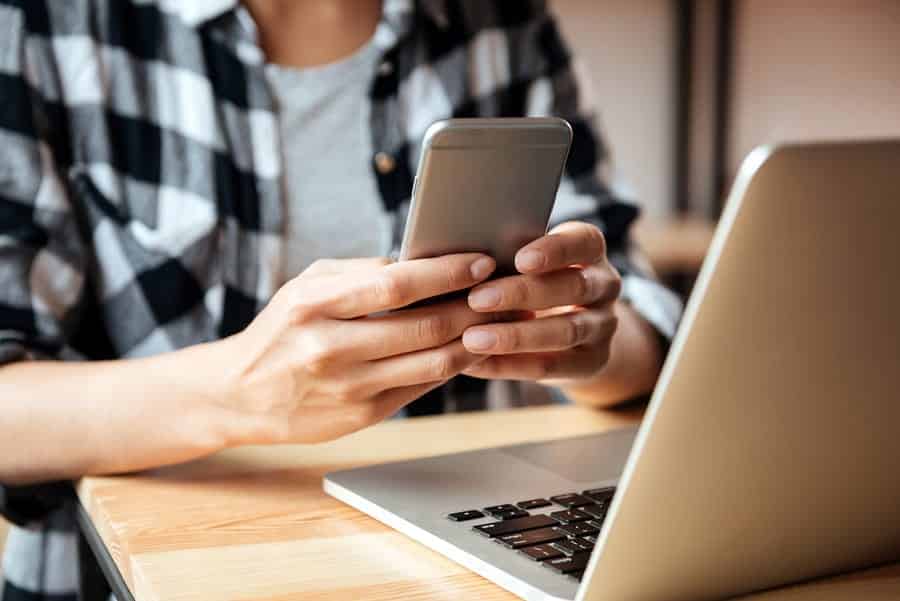 A photo of a woman checking something on her iPhone in between working on her laptop.