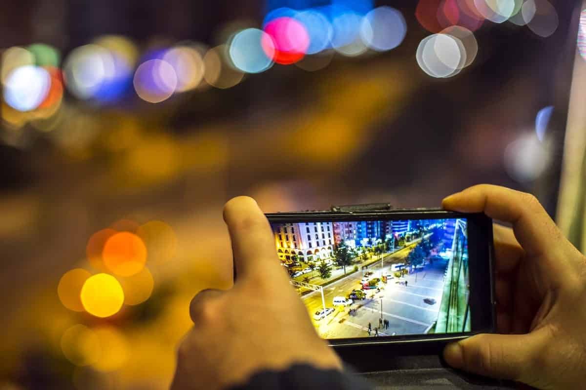 A photo of a person taking a picture with his smartphone of a brightly lit street at night.