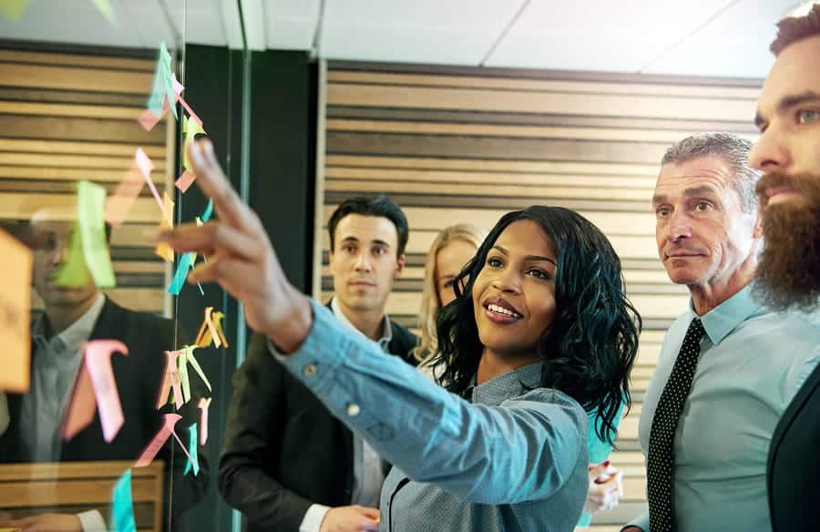 A photo of a woman showing a work group a series of multi-colored sticky notes on a window.