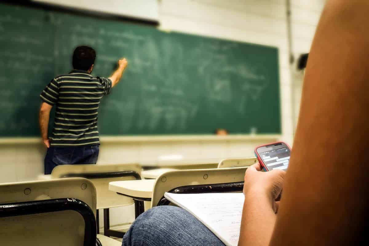 A photo of a student on their phone will the teacher explains something at the chalkboard.