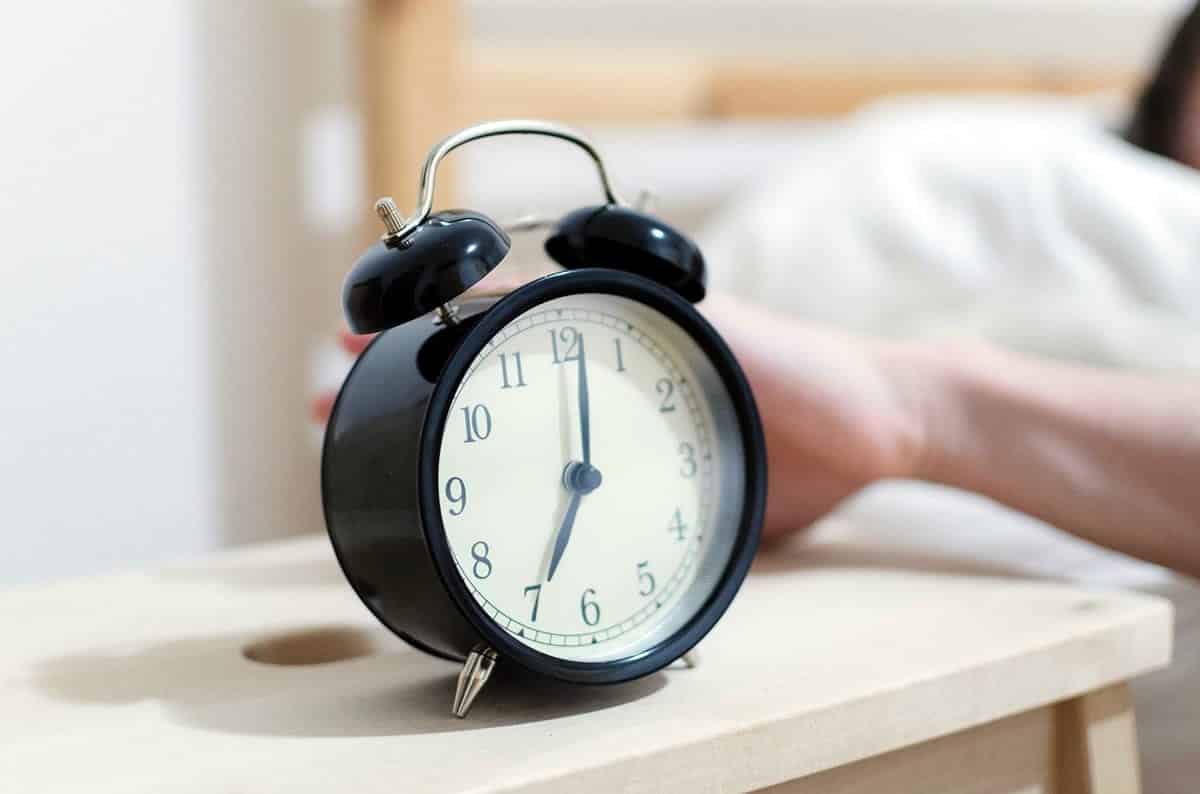 An image of a person in bed with their hand behind an alarm clock.