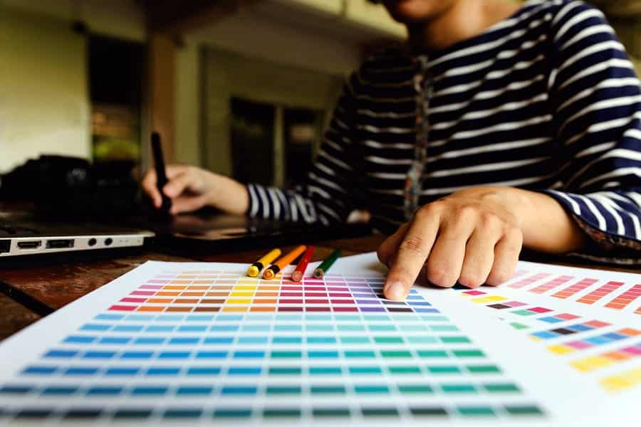 A photo of a designer drawing on a tablet while referencing a sheet full of color swatches.