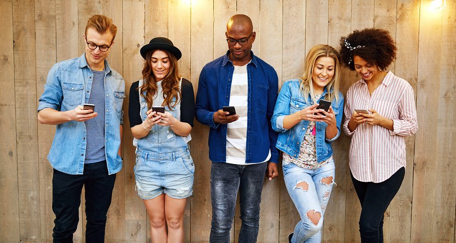 A photo of five people on their smartphones leaning against a fence.