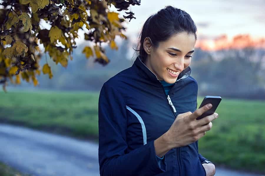 A photo of a woman taking a walk in the early morning, smiling at something on her smartphone.