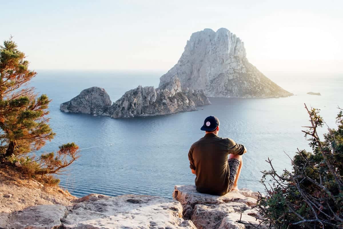 An image of a man on vacation, sitting on a rock by the sea.  