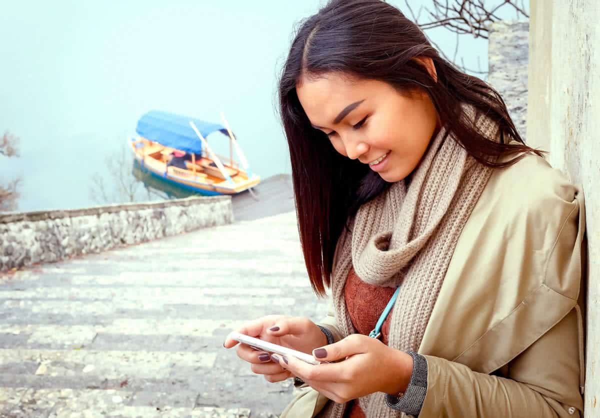 A photo of a woman leaning against a wall and looking at her smartphone.