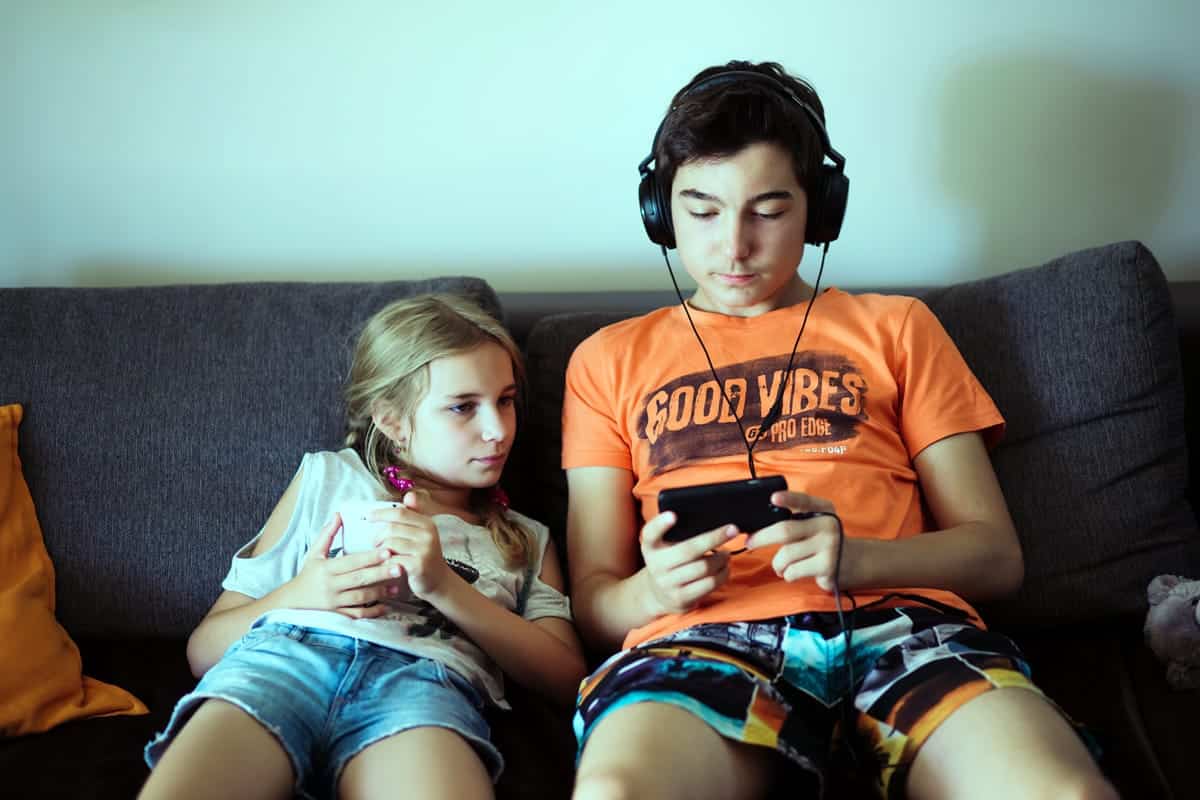 A photo of a brother and sister sitting on the couch, playing with their smartphones.