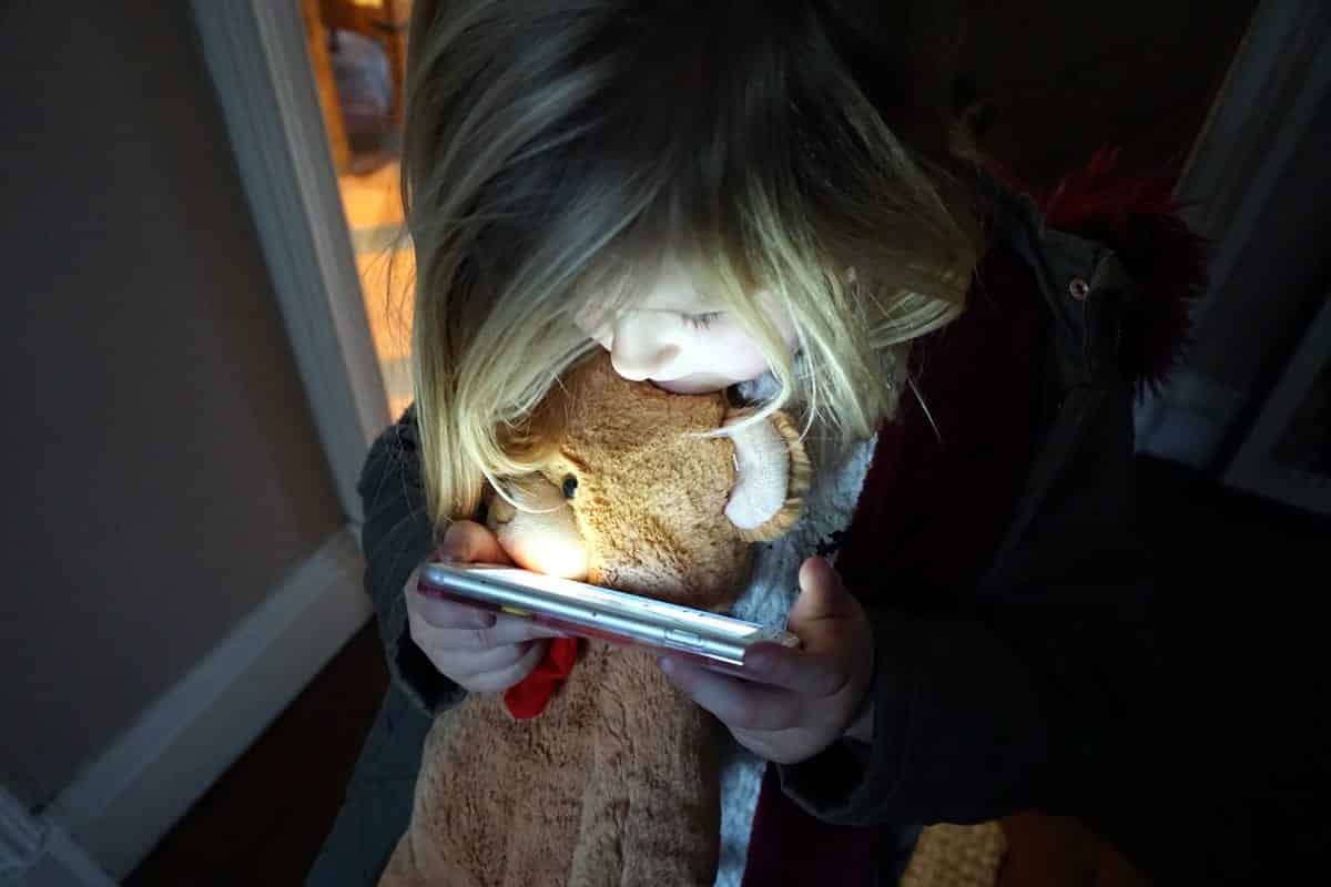 A photo of a little girl holding a teddy bear and watching something on a smartphone.