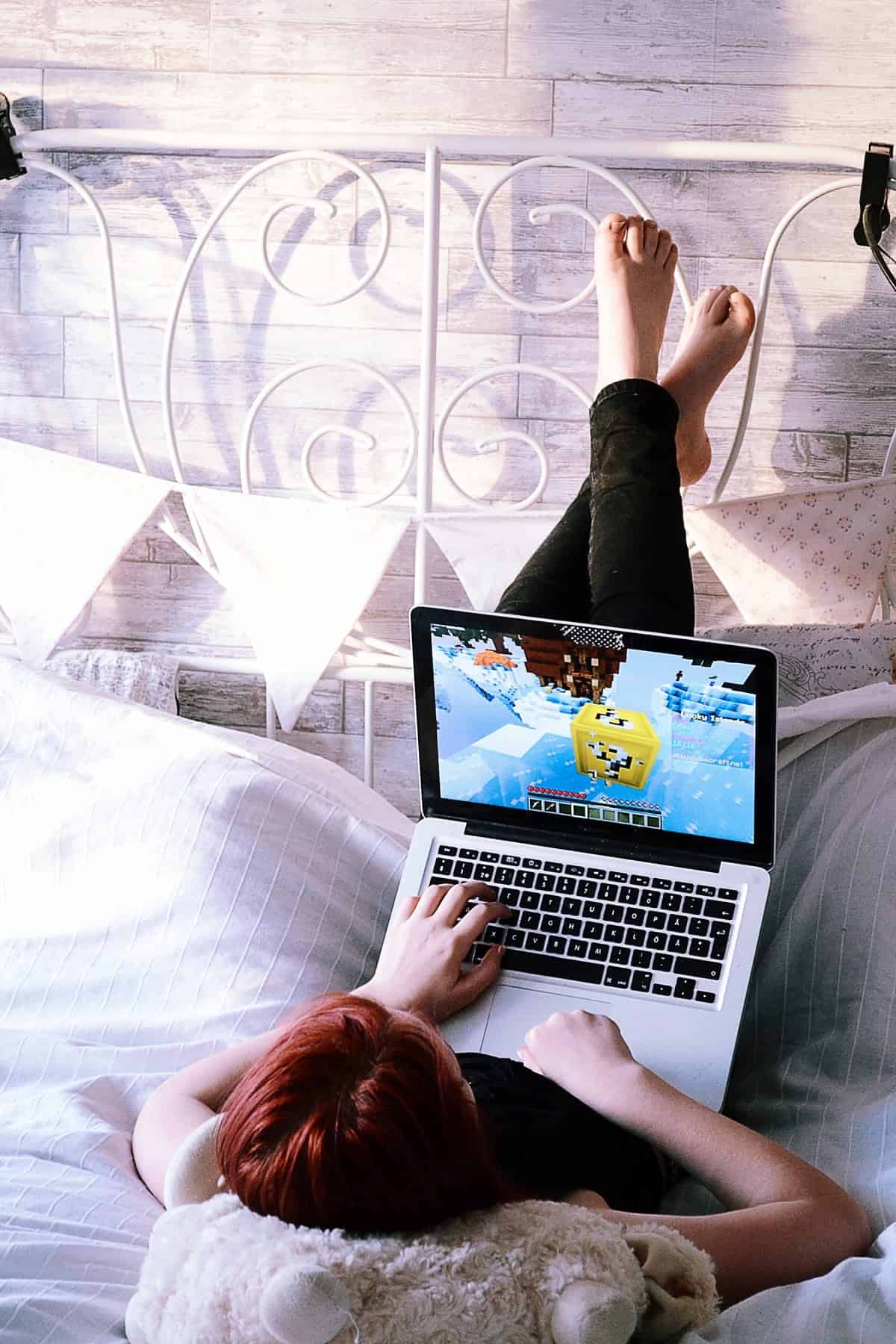 A photo of a woman lying with her feet up against a headboard, playing games on a laptop.