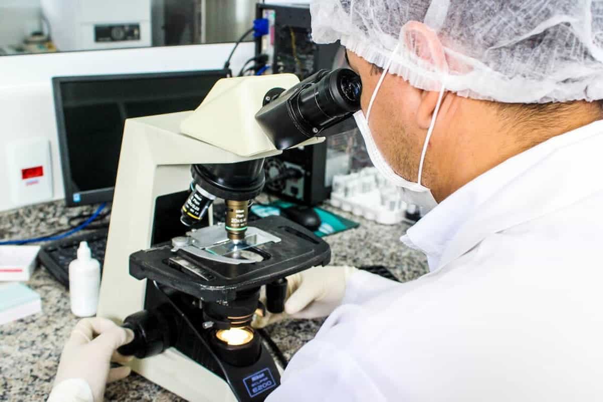 A photo of a man wearing a protective hair net and mask looking through a microscope.