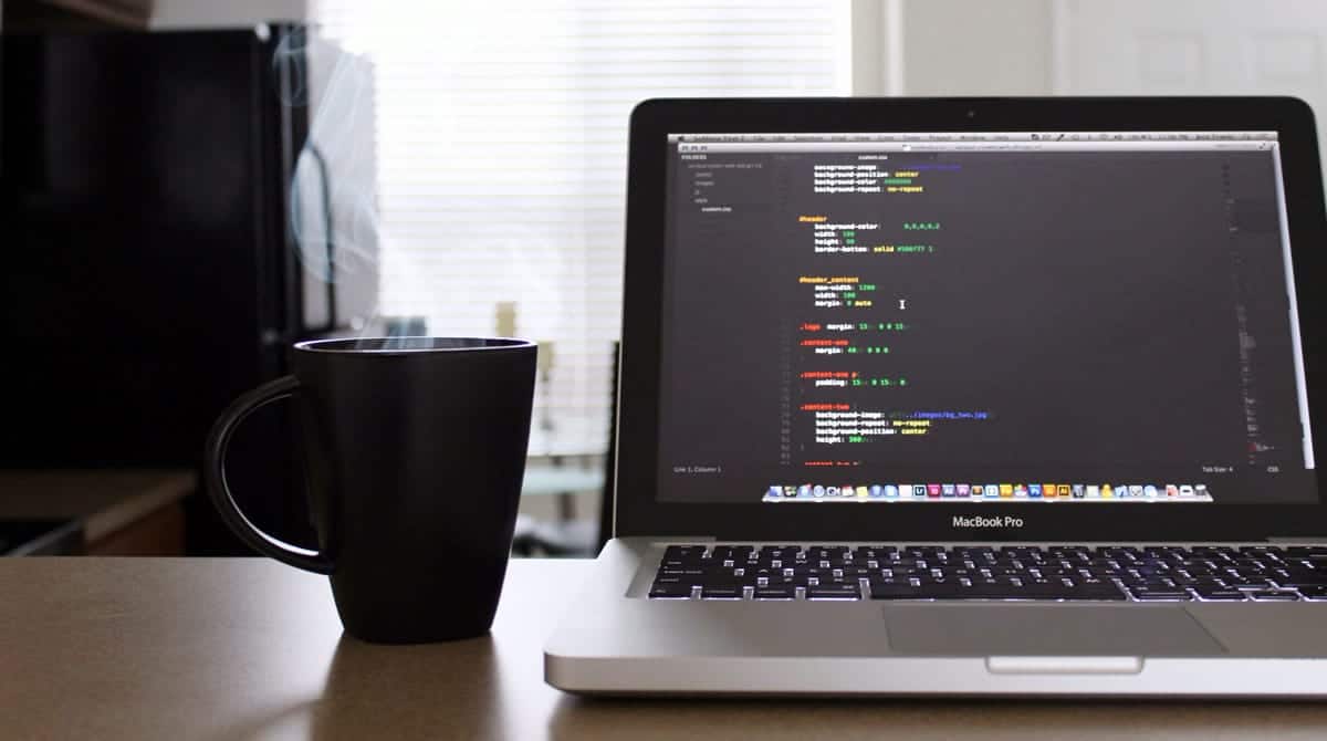 A photo of a steaming cup of coffee next to a laptop displaying lines of code.