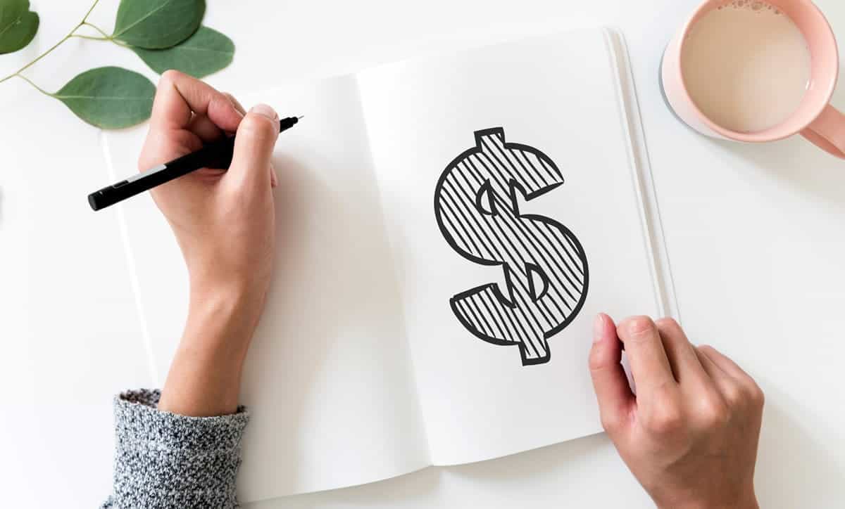 A person drawing a dollar sign in a notebook on a table with a mug and leaves.