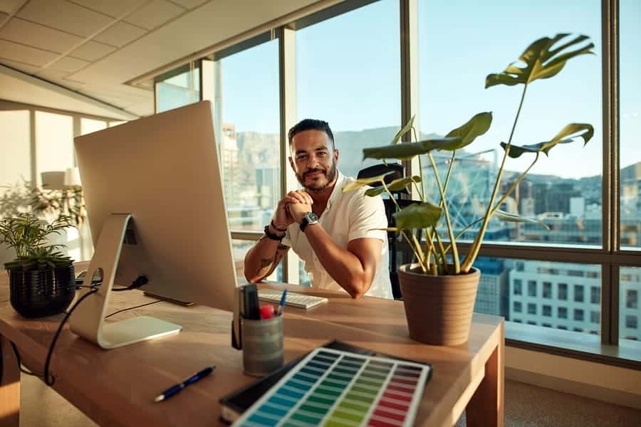 A photo of a smiling entrepreneur sitting in his office with floor to ceiling windows.