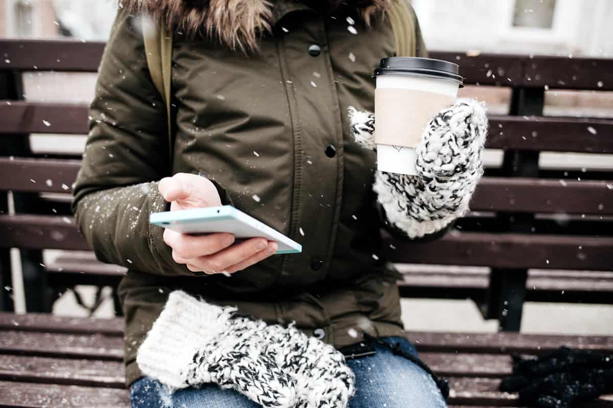 A photo of a woman sitting on a bench in the snow, holding coffee in one hand and her smartphone in the other.