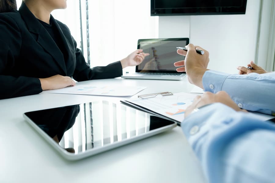 A photo of people sitting at a table during a meeting and discussing a chart on a tablet.