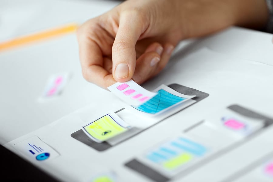 A close-up photo of a person placing pieces of paper onto a drawing of a mobile phone, demonstrating mobile app design decisions.