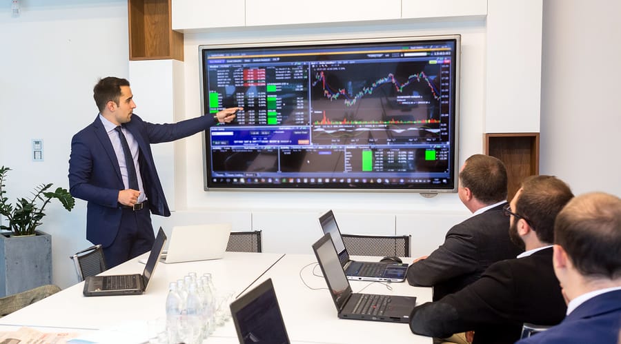 A photo of a man giving a presentation with color-coded graphs on a large screen.