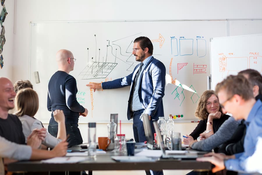 A photo of two men looking at drawings on a whiteboard while the rest of the product management team discusses their own progress.