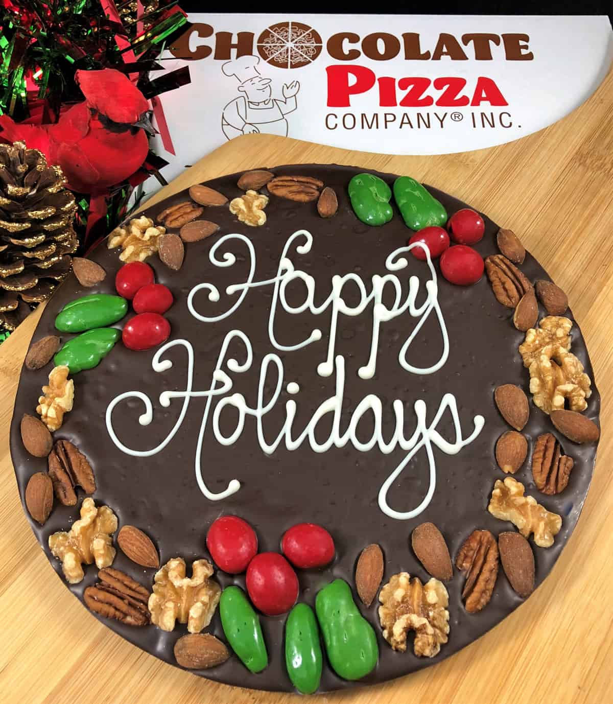 A photo of a chocolate pizza with “Happy Holidays” written on it and decorated with a border made of pecans, walnuts, and almonds.