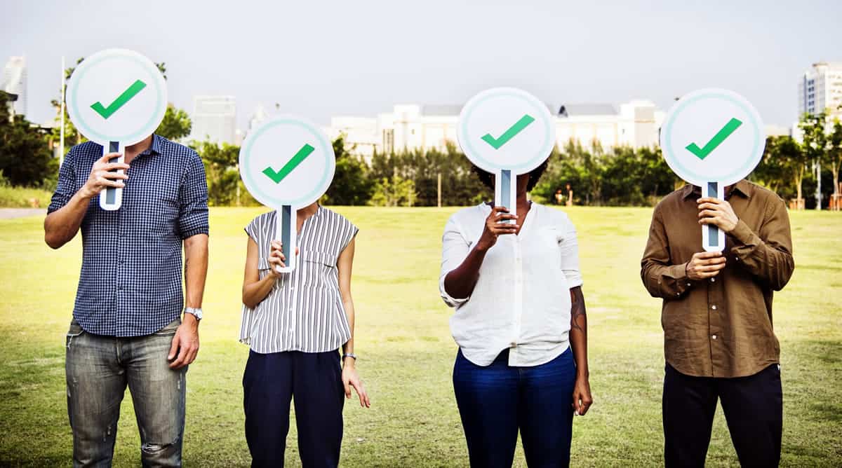 Four people standing outdoors with signs with green check marks over their faces.