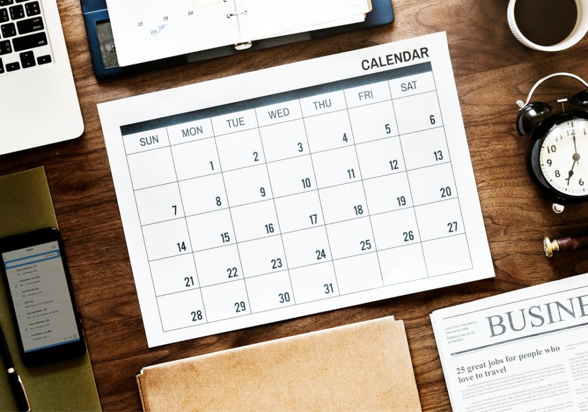 A calendar, computer, clock, and day planner on a table.