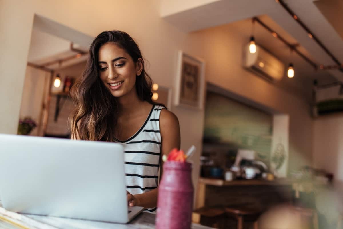 A photo of a woman working on her computer in the morning with a smoothie on her desk.