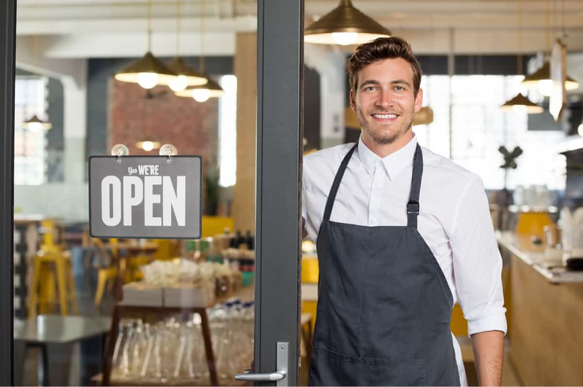 A photo of a man standing next to an “open” sign with a proud smile on his face.