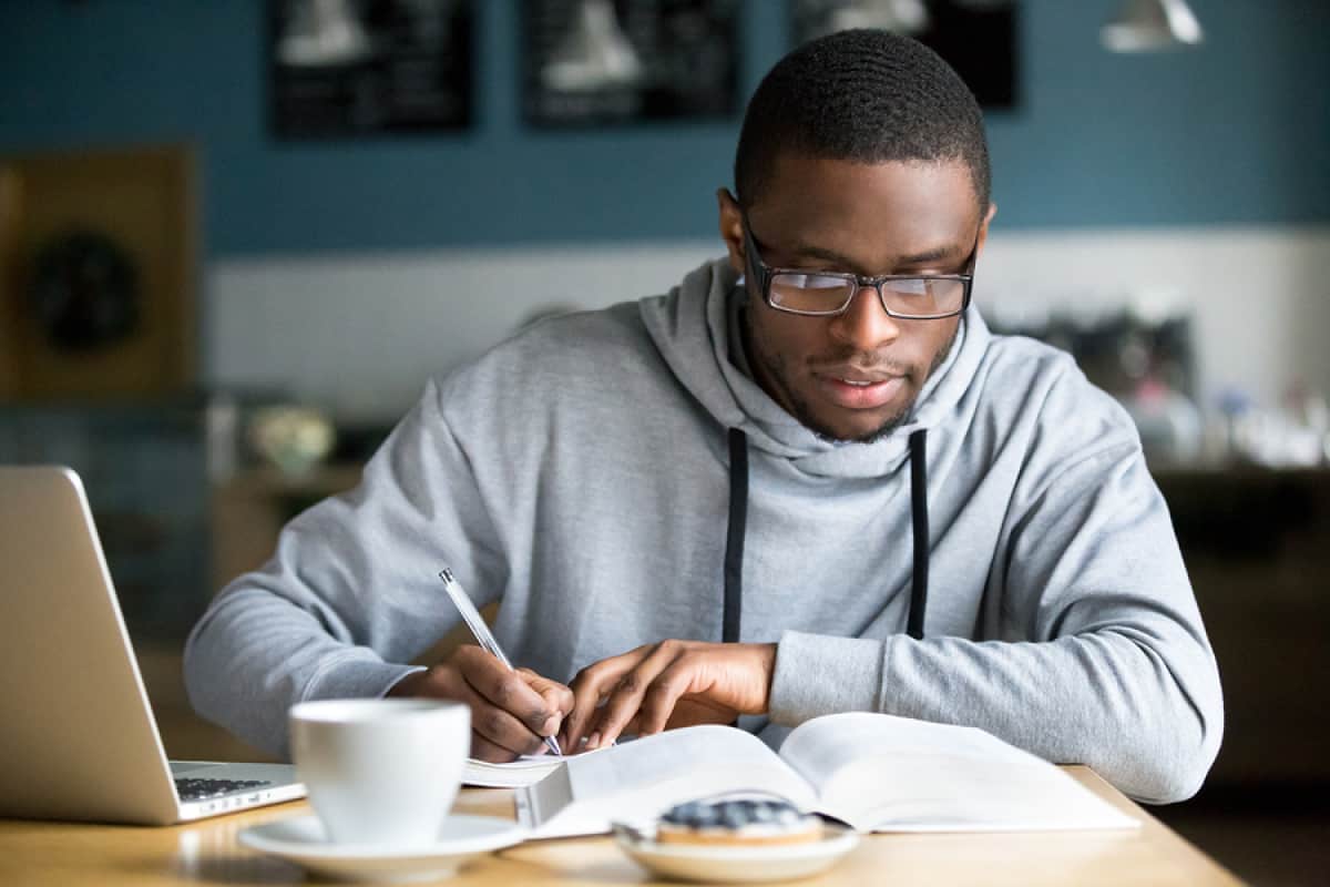 A photo of a man taking notes out of a book over morning coffee and a donut.