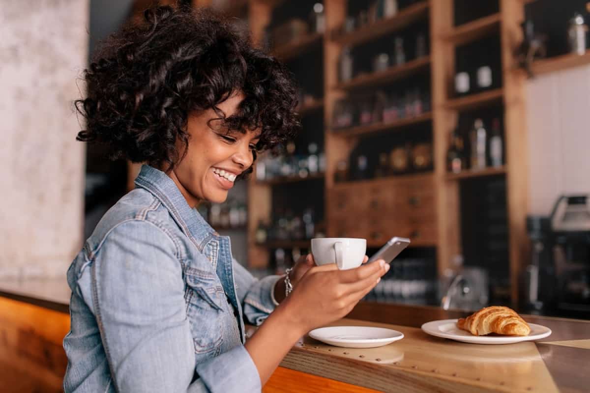 A photo of a happy woman in a coffee shop, reading a message on her smartphone.