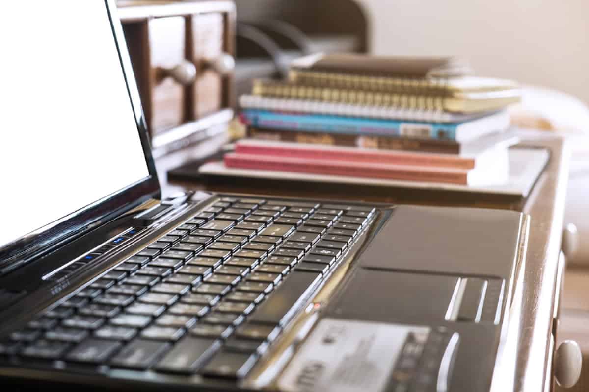 A photo of a laptop sitting on a desk with a stack of books and notebooks in the background.