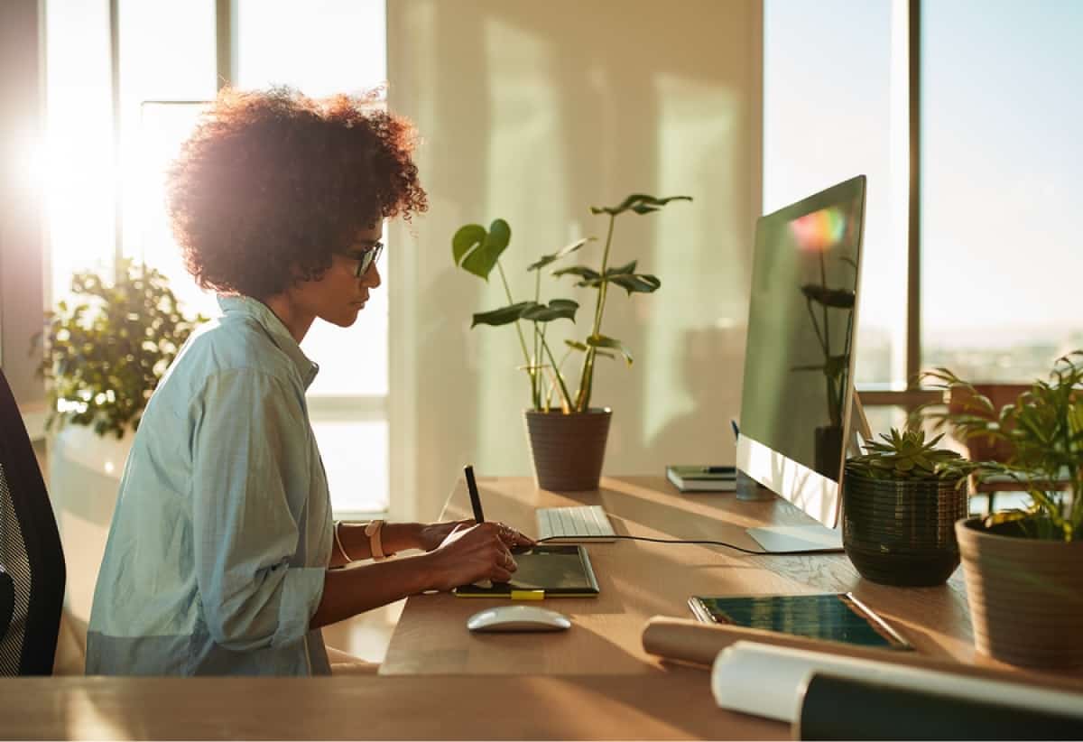 A photo of a young woman working at her desk with the sun shining brightly through the window.