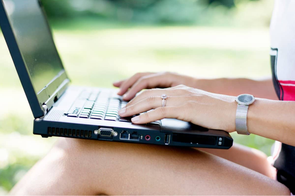 A photo of a laptop being balanced on a woman’s knees as she works in a park.