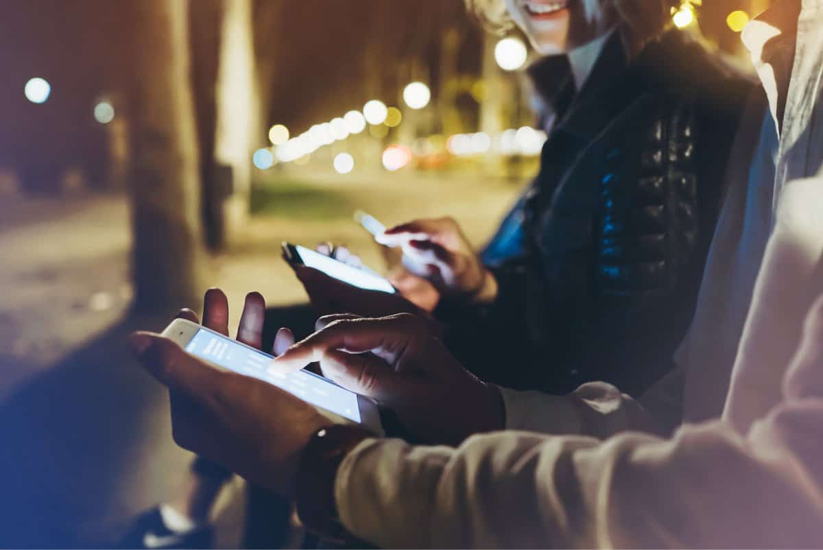 A photo of three friends on a street after dark, checking their smartphones.