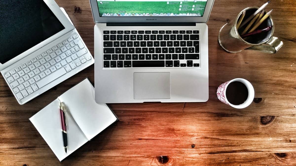 A photo of someone’s desk with a tablet, laptop, notepad, pencil cup, and coffee mug.