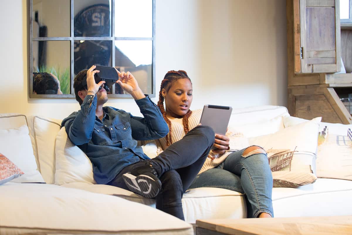 A photo of a couple sitting on the couch, playing with a tablet and a virtual reality headset.