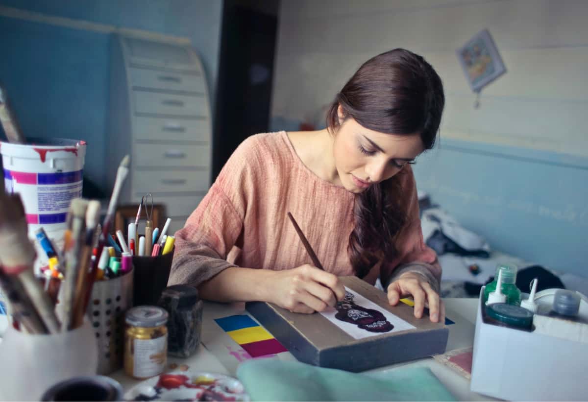 A woman drawing at a desk surrounding by art supplies.