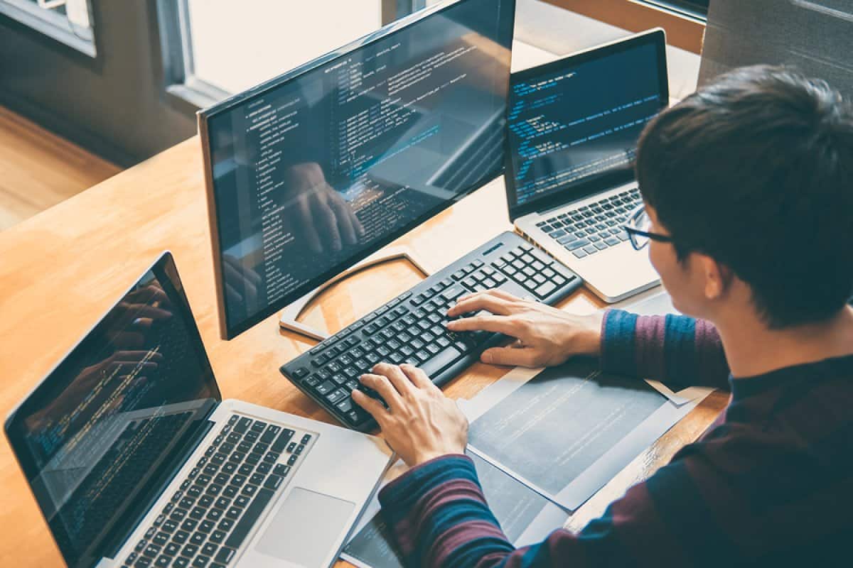 A photo of a programmer sitting at his desk with three computers in front of him, each displaying lines of code.