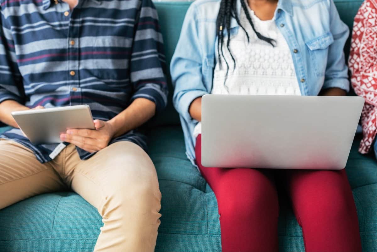 Two people sitting on a couch, one taking notes on their computer and the other testing an app on a tablet.