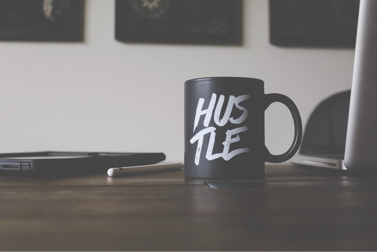 A photo of a coffee mug that says “HUSTLE” sitting on a desk with a laptop and a tablet.