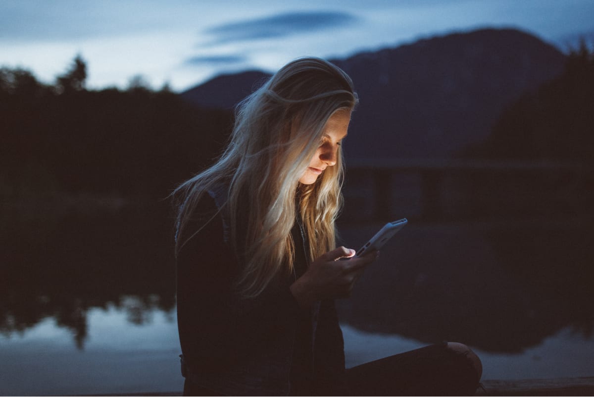 A photo of a woman sitting by the water, checking a mobile app on her smartphone.