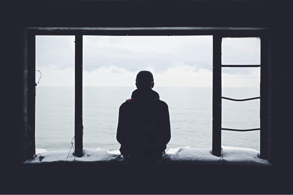 A photo of a person sitting on a snowy windowsill, overlooking a body of water.