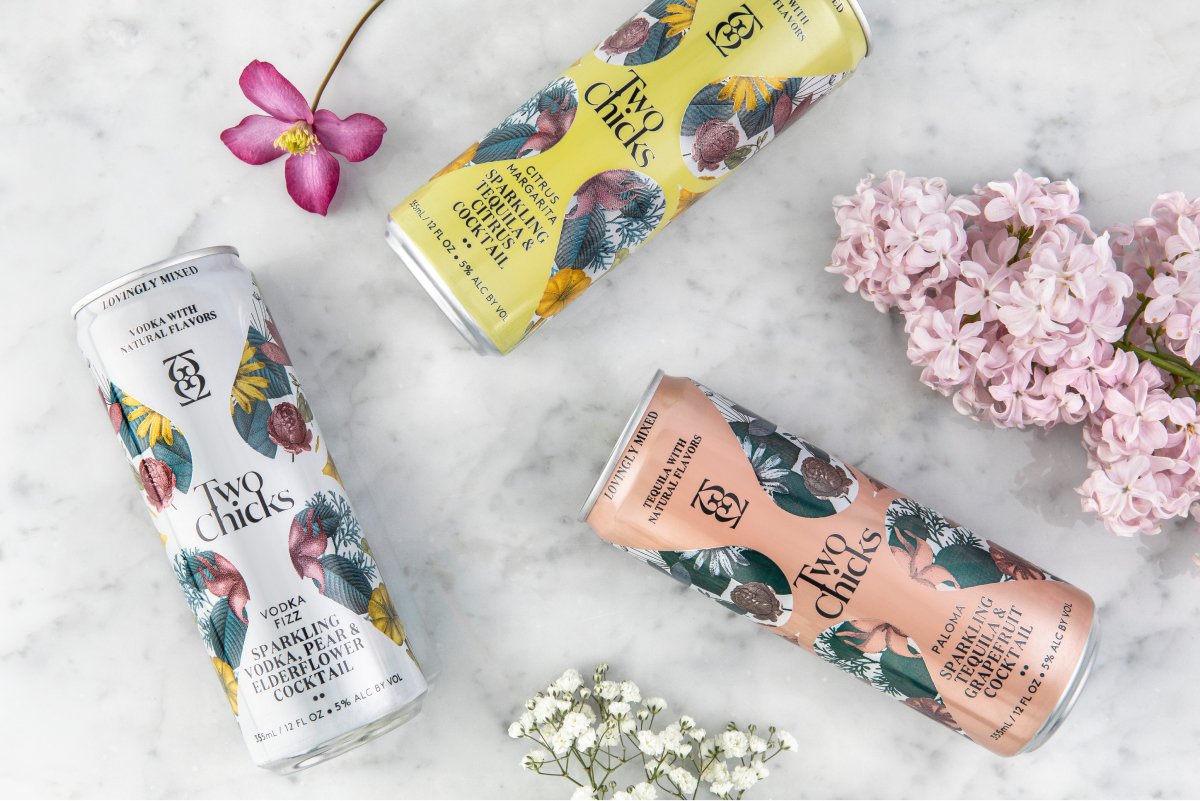 A photo of Two Chicks Cocktails cans laying on a marble surface among cut flowers.