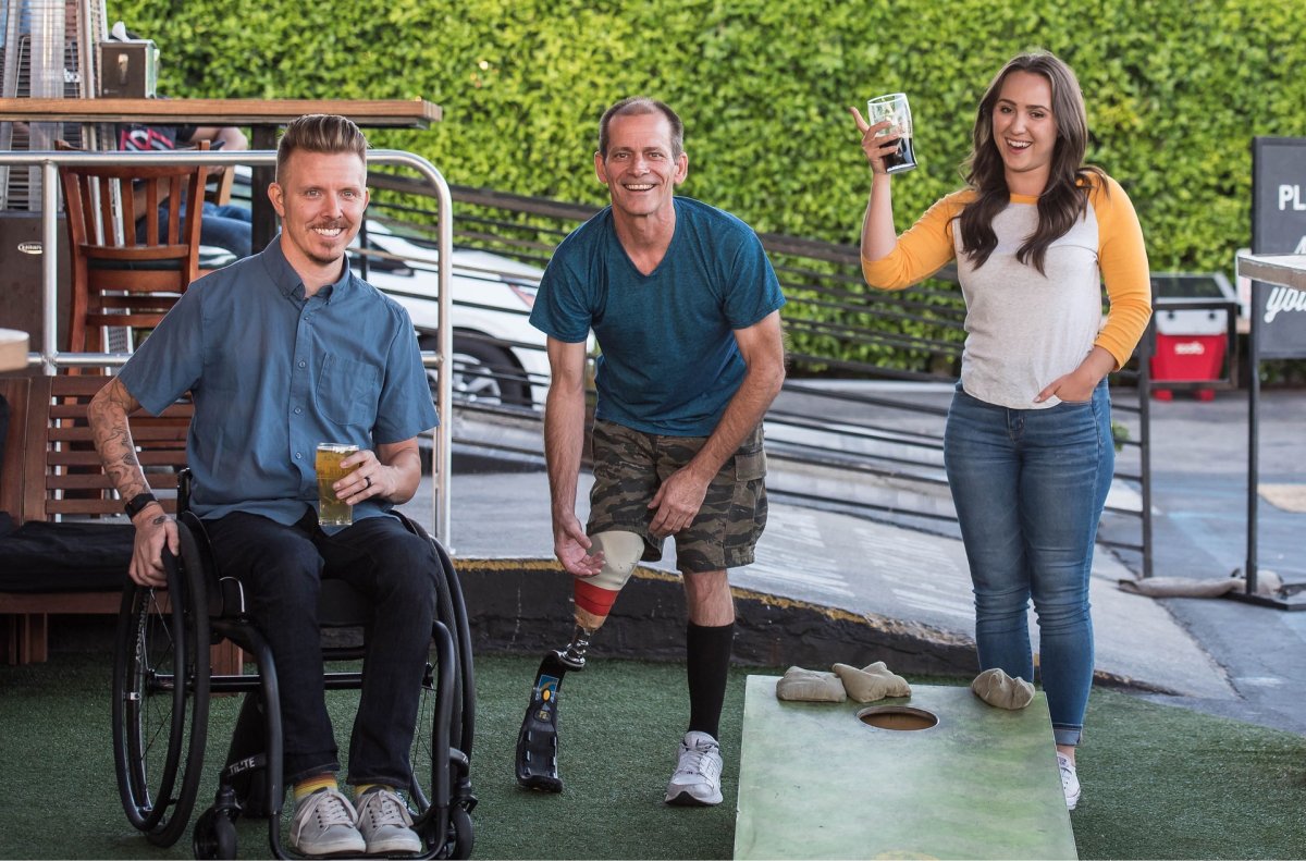 A man in a wheelchair, a man with a prosthetic leg, and a woman drinking beer and playing cornhole. 